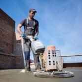 Full length of man laborer using troweling machine while screeding floor in new building under blue sky. Male worker finishing concrete surface with floor screed grinder machine at construction site.
