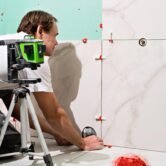 Professional master man laying ceramic tiles on wall in bathroom. Portrait of an experienced repairman laying large size porcelain tiles.Construction worker checking tile installation with laser level